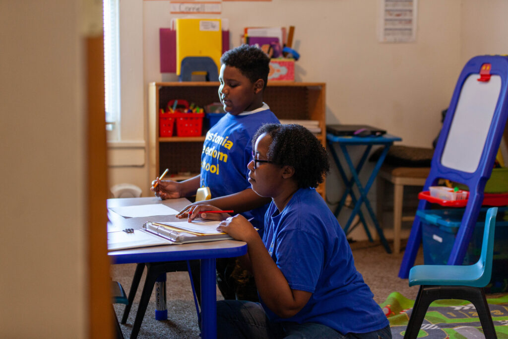 looking from th door into a classroom, we see a black student around age 7 sitting at a short table with a black woman sitting on the floor beside him. They are looking at papers on the desk.